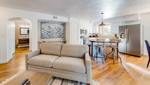 Ein modernes Wohnzimmer mit einer Couch, einem Essbereich mit Tisch und Stühlen, einer Küche mit Geräten und einem großen abstrakten Gemälde an der Wand.