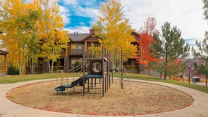 Ein Spielplatz mit Rutsche und Klettergerüsten, umgeben von Bäumen mit Herbstlaub, in einem Wohngebiet mit Häusern im Hintergrund.