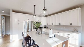 A modern kitchen with white cabinetry, a central island, stainless steel appliances, pendant lighting, and a potted plant. - Bright Spacious & Modern Condo with fireplace and gas grill (Heber City)