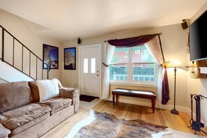 Ein Wohnzimmer mit einer Couch, einer Bank, einem Fenster mit Vorhängen, einer Lampe und einem Fernseher.