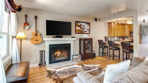 Una sala de estar con chimenea, televisor, una guitarra y una cocina al fondo.