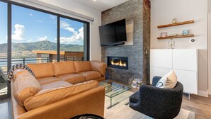 A modern living room with a leather sofa, a glass coffee table, a fireplace, a flat-screen TV, and a view of mountains. - Townhouse in Hideout with hot tub (Hideout)