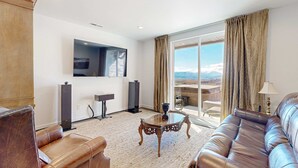 A living room with a sofa, a coffee table, a TV mounted on the wall, and a large window with curtains. - Stunning townhome with  unobstructed views, amenities for the health conscious (Heber City)