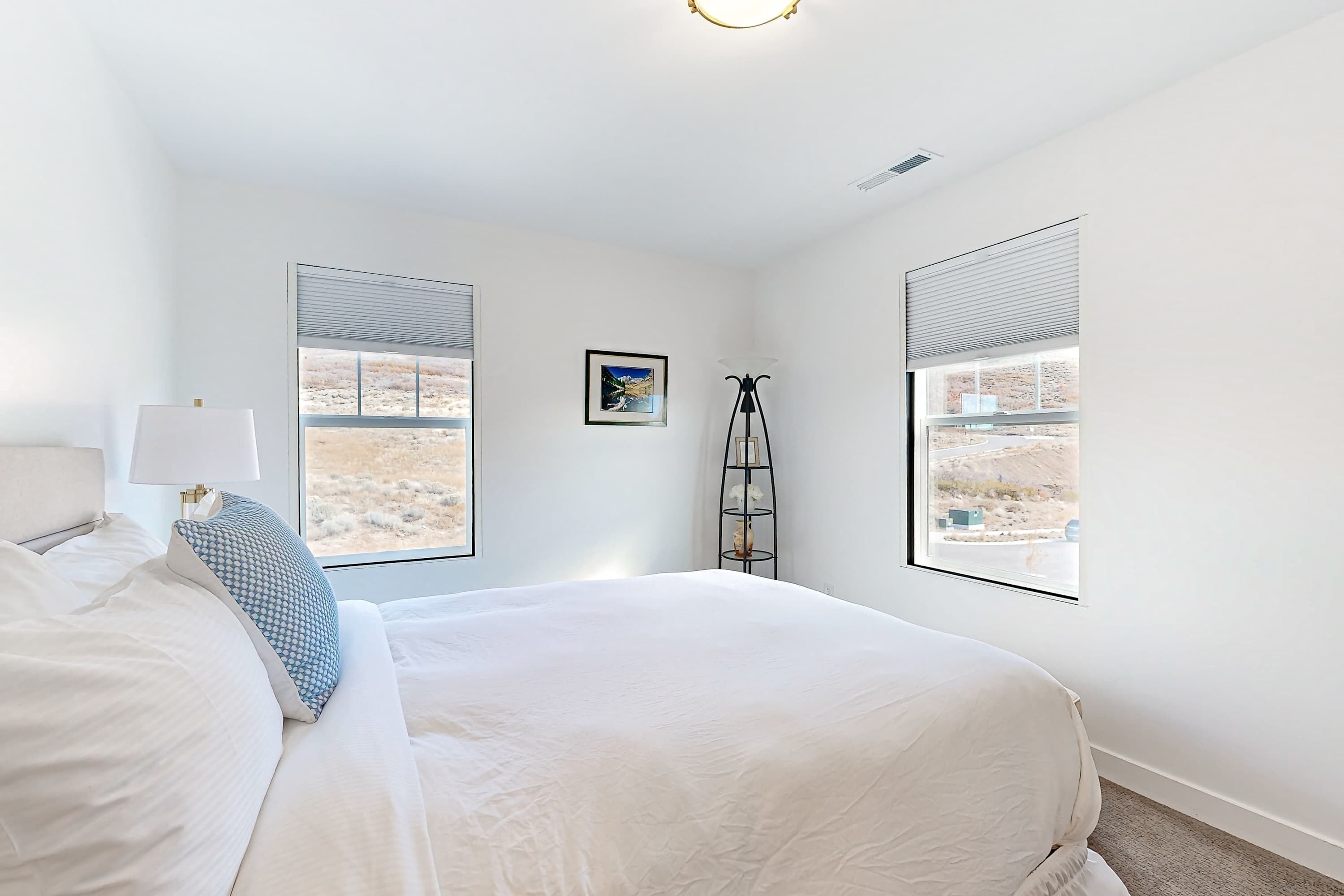 A bedroom with a large bed, two windows with blinds, a floor lamp, and a picture on the wall.