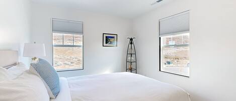 A bedroom with a large bed, two windows with blinds, a floor lamp, and a picture on the wall.