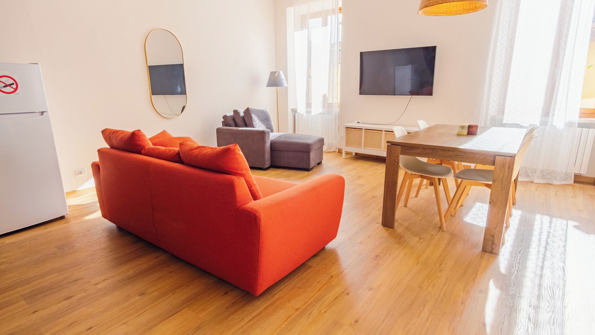 Una sala moderna con un sofá rojo, un sillón gris, una mesa de comedor de madera con sillas, un televisor de pantalla plana y un refrigerador.