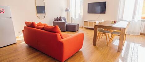 Una sala moderna con un sofá rojo, un sillón gris, una mesa de comedor de madera con sillas, un televisor de pantalla plana y un refrigerador.
