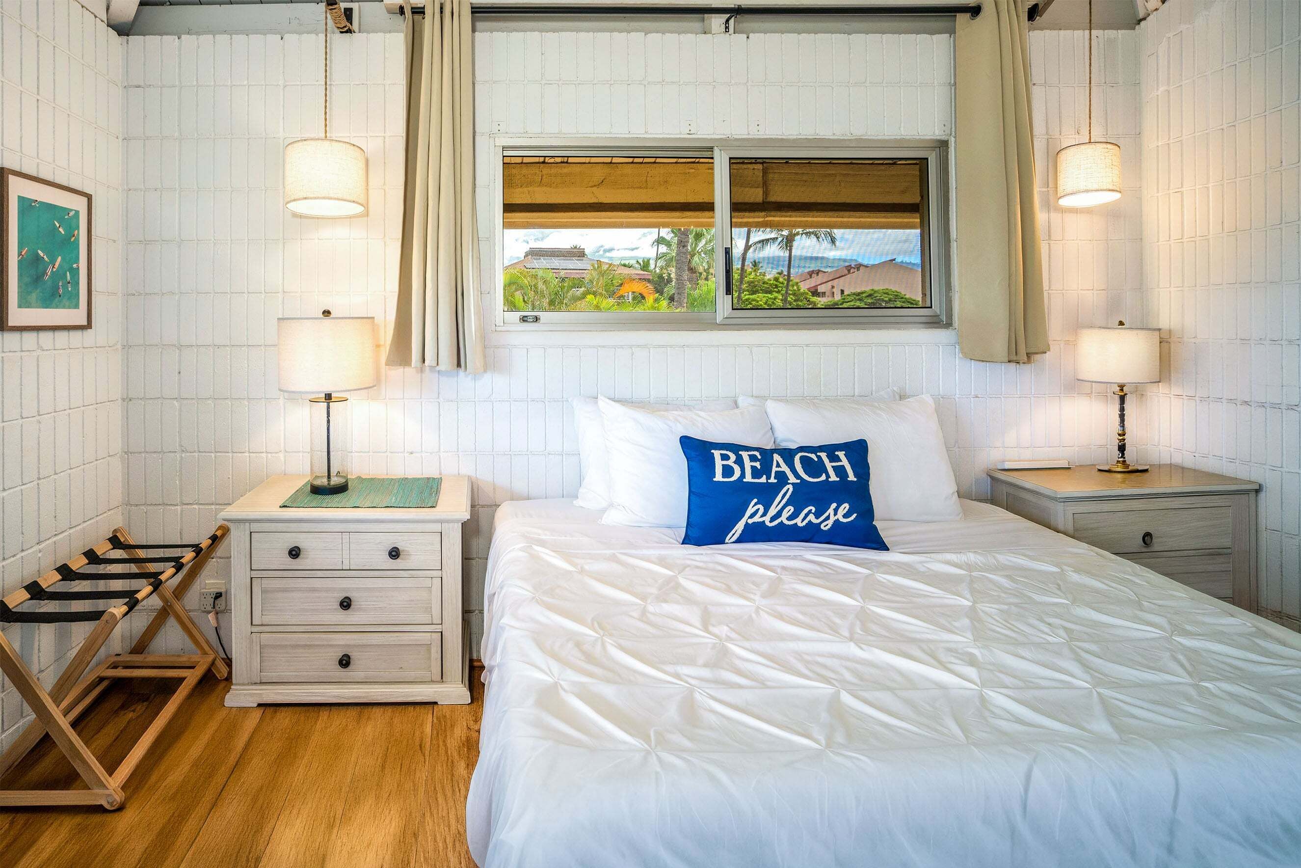 Een net opgemaakt bed met een kussen waarop 'STRAND alstublieft' staat, een houten droogrek en twee bedlampjes.