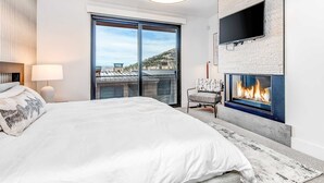 A modern hotel room with a large bed, a fireplace, a TV mounted on the wall, and a balcony with a city view. - Elegant townhome with mountain view, private hot tub, fireplace & lovely decor (Park City)