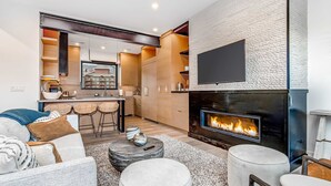 A modern living room with a fireplace, a kitchen in the background, and a dining area with bar stools. - Elegant townhome with mountain view, private hot tub, fireplace & lovely decor (Park City)