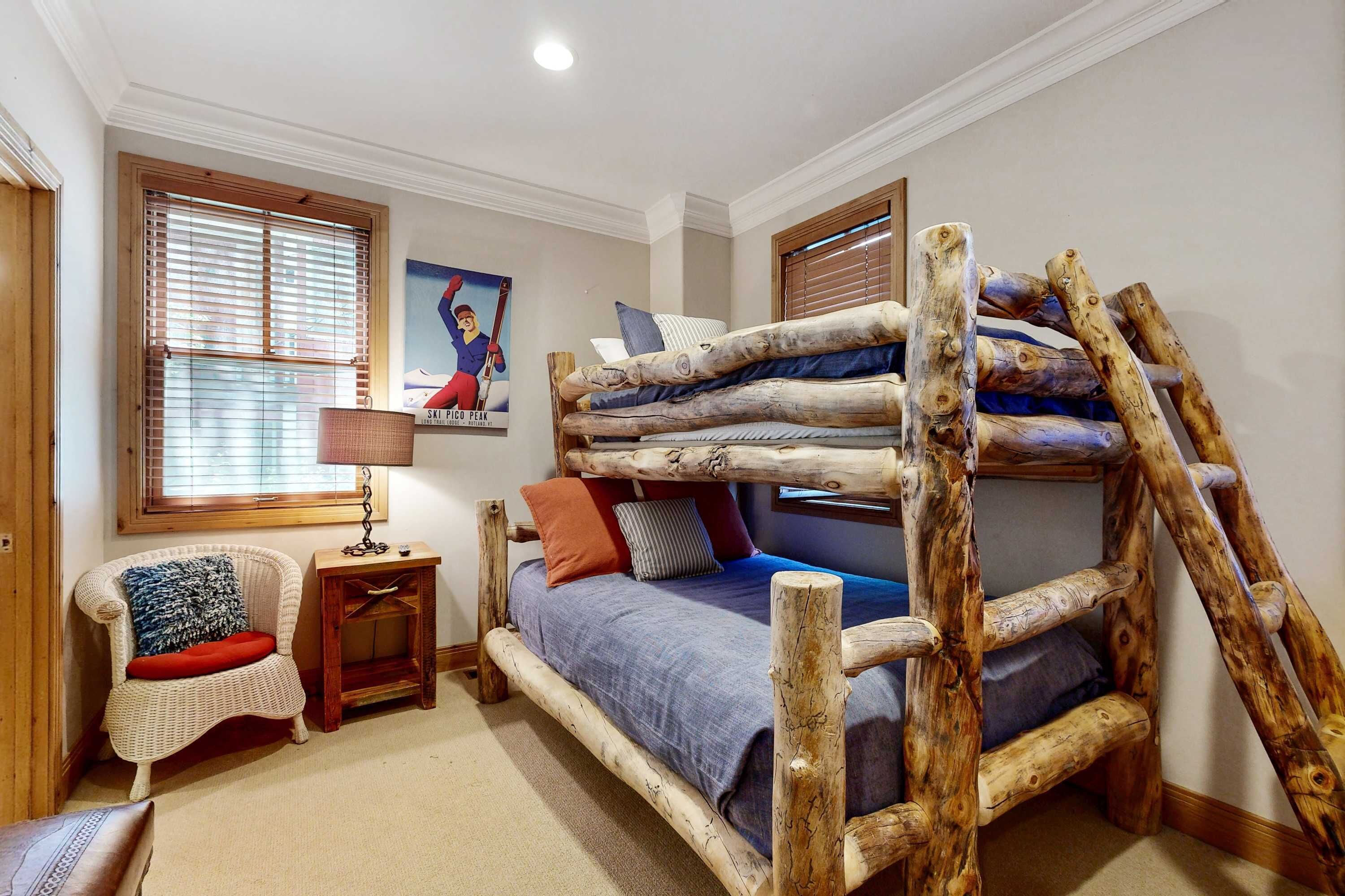Una cama litera con estructura de madera, una silla con cojín, una mesita con lámpara y un cartel en la pared.