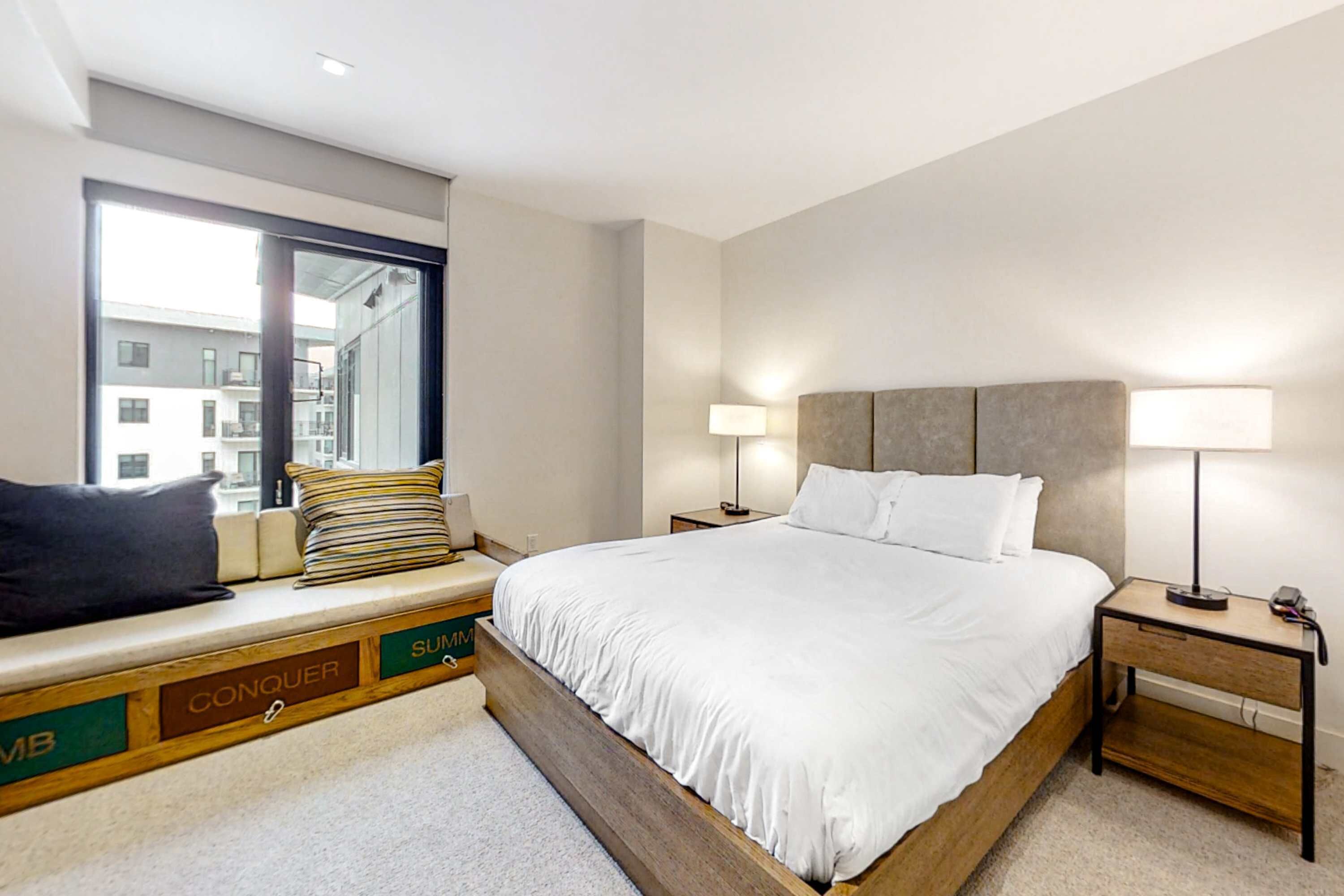 A modern hotel room with a large bed, bedside lamps, a window seat with cushions, and a view of buildings outside.