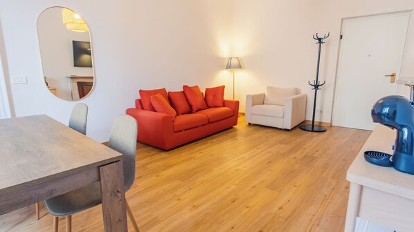 Ein modernes Wohnzimmer mit Holzboden, einem roten Sofa, einem weißen Sessel, einem Esstisch mit Stühlen, einem Kleiderständer und einem an der Wand befestigten Fernseher.