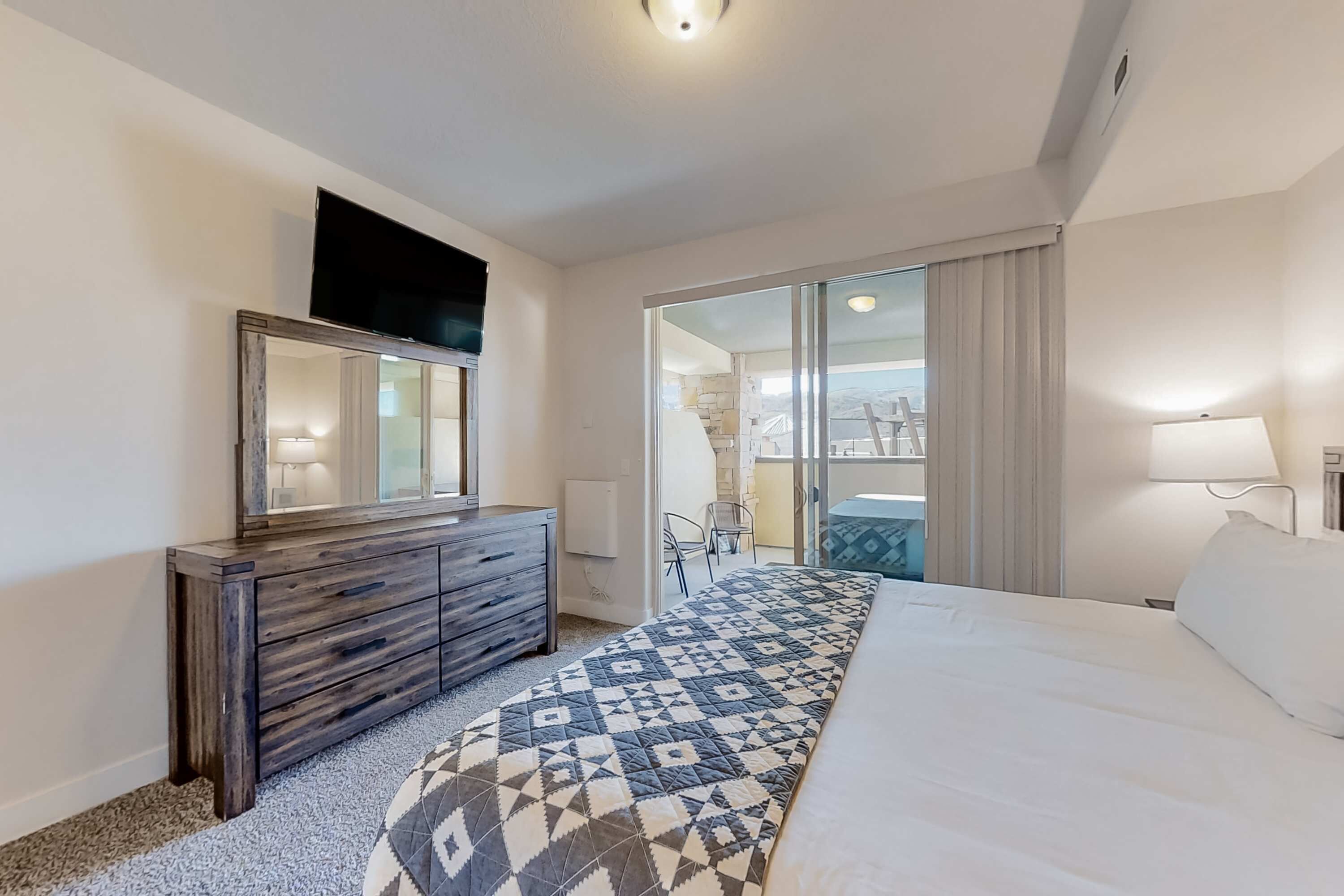 A hotel room with a bed, a dresser, a TV, a balcony with a view, and a window with curtains.
