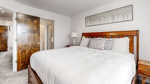 Une chambre à coucher avec un grand lit, une tête de lit en bois, du linge de lit blanc, une table de chevet avec une lampe et une porte donnant sur une autre pièce.