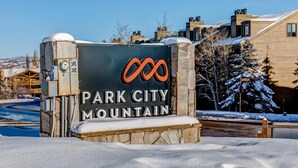 Un panneau indiquant l’entrée de Park City Mountain, avec une base en pierre et une partie supérieure en métal, entouré de neige et de bâtiments.