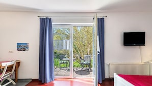 Una habitación con cama, televisor, mesa y sillas, y vistas a través de un ventanal a un espacio exterior con mesa y sillas.