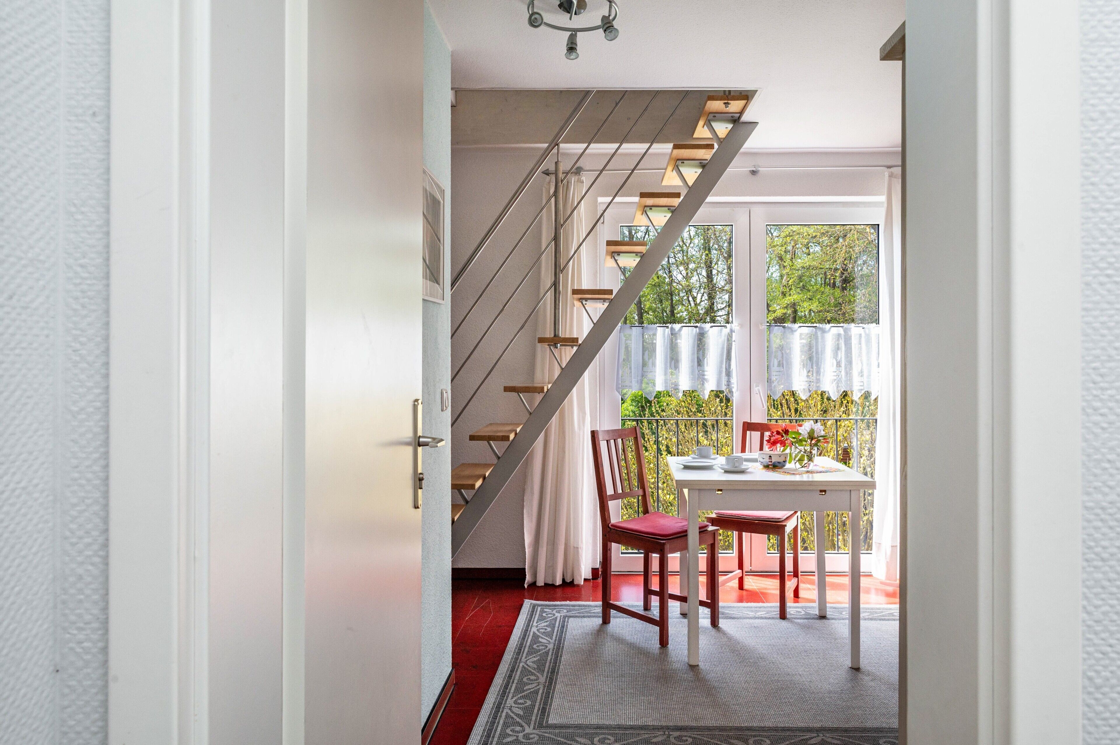 Eine Treppe führt zu einem Balkon mit Tisch und Stühlen, einem Fenster mit Vorhängen und einem gemusterten Teppich.