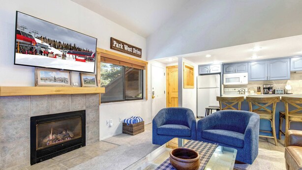 A living room with a fireplace, a television mounted above it, and a kitchen area visible through an open door. - Two Lofts, Mountain Views, Gas Fireplace, Shared Seasonal Hot Tub & Pools (Park City)