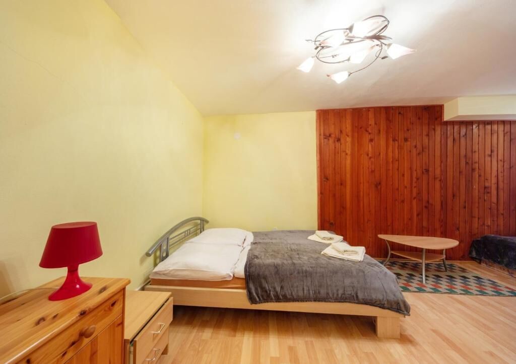 Une chambre à coucher avec un lit, une lampe rouge, une commode en bois, une petite table ronde et un luminaire au plafond.