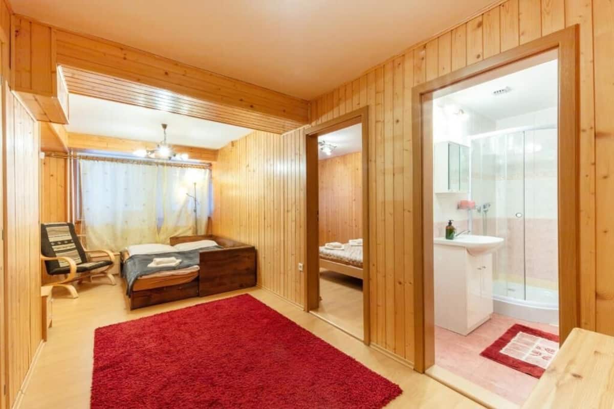 A wooden-paneled room with a bed, a red rug, a chair, and a bathroom visible through a glass door.