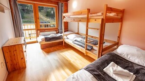 Een slaapkamer met stapelbed, houten vloer, een raam met gordijnen en een klein houten kastje.