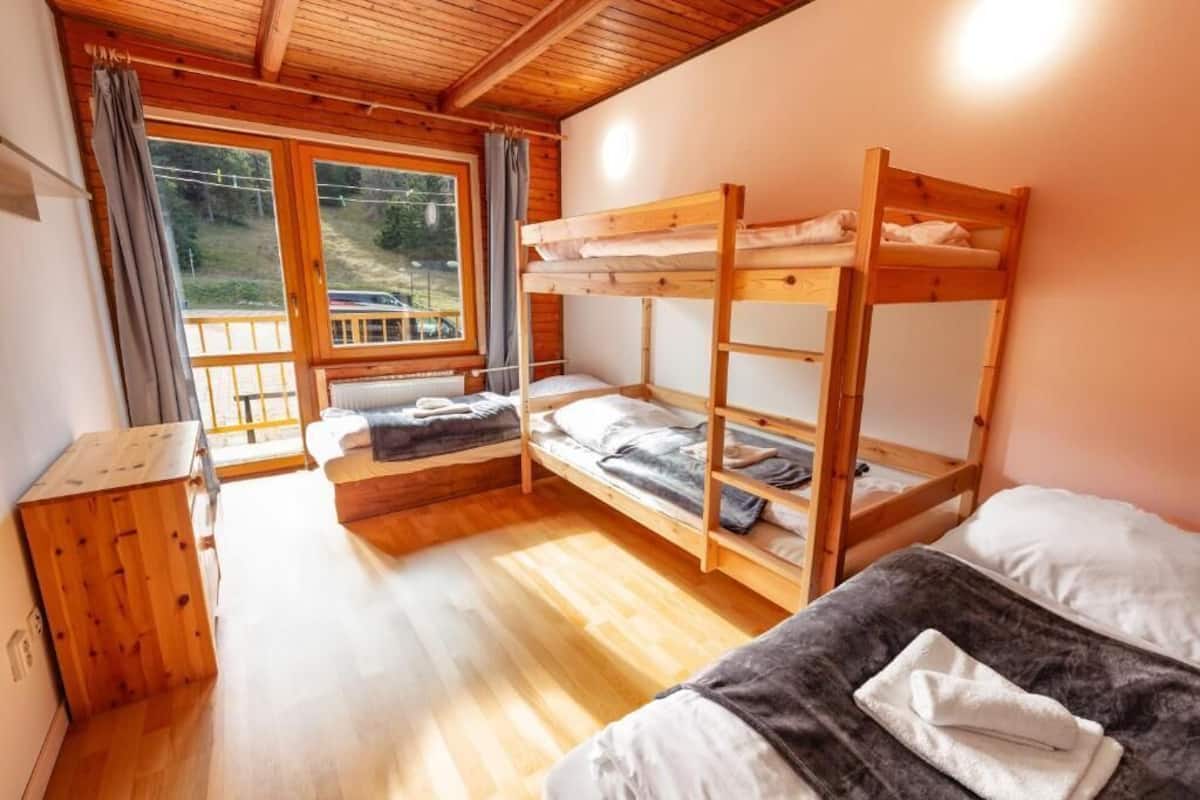 A bunk bed room with a wooden floor, a window with curtains, and a small wooden cabinet.