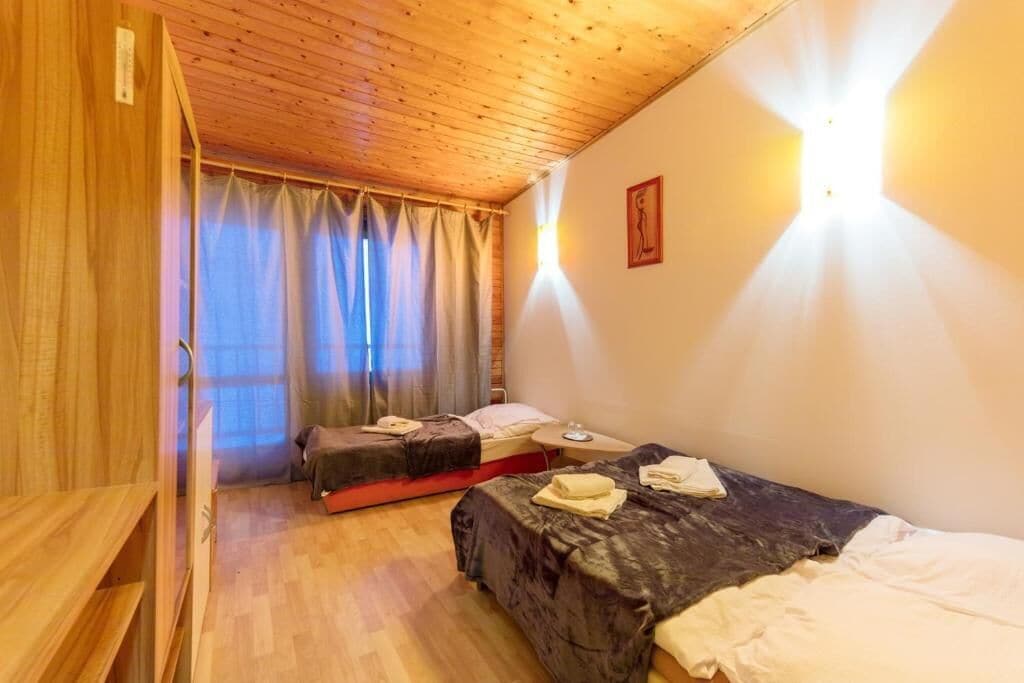A room with wooden ceiling, two beds, a bedside table, a window with curtains, and a wall-mounted light.