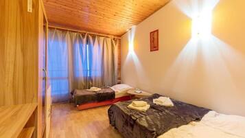 A room with wooden ceiling, two beds, a bedside table, a window with curtains, and a wall-mounted light.