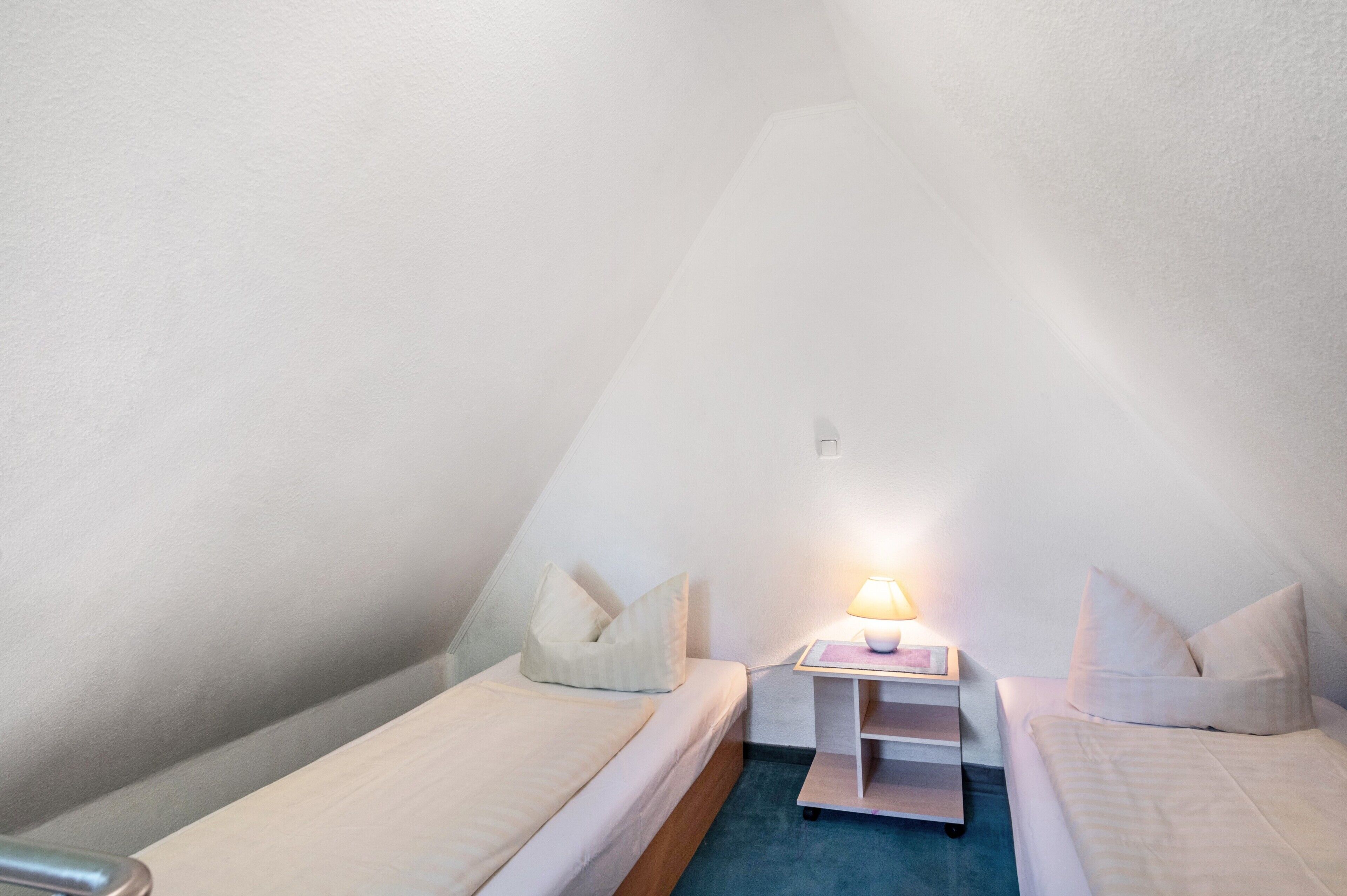 Una stanza piccola e triangolare con due letti singoli, un comodino con lampada e una mensola a parete.