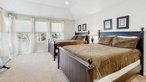 Une chambre avec deux lits, une grande fenêtre avec des rideaux, une table de chevet avec une lampe et des tableaux encadrés au mur.