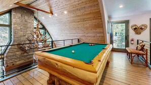 A wooden pool table in a room with a stone wall, wooden ceiling, and large windows.