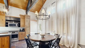 Una cocina con una mesa redonda de comedor, armarios de madera, una araña y un gran ventanal con cortinas.