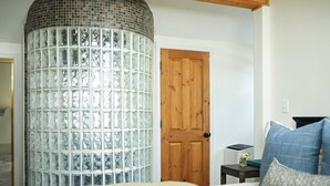 Un dormitorio con cama, una puerta de madera, una mesita con lámpara y una pared de bloques de vidrio.