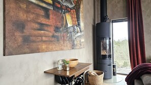 A room with a large abstract painting, a black wood-burning stove, a wooden table with a bowl and a lantern, and a view through a window with curtains. - Villa with Pool and Sea View on Orust | SE09168 (Varekil)