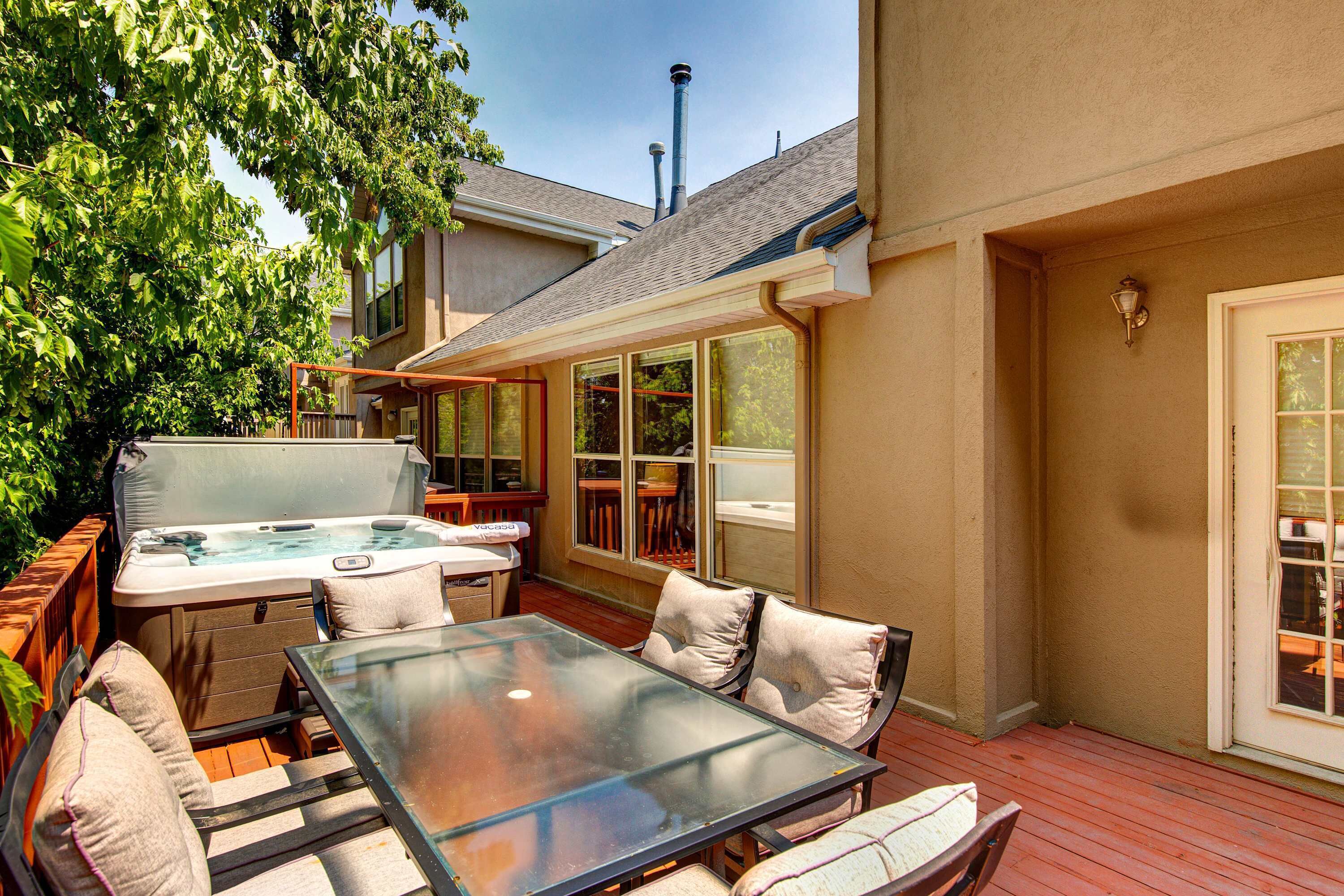 An outdoor patio with a glass table, cushioned chairs, a hot tub, and a wooden deck.