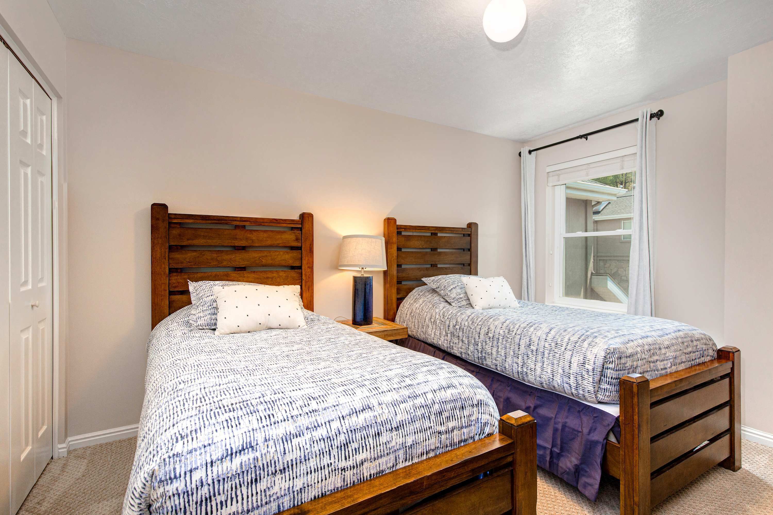 Un dormitorio con dos camas de madera, una mesita de noche con lámpara, una ventana con cortinas y una puerta.