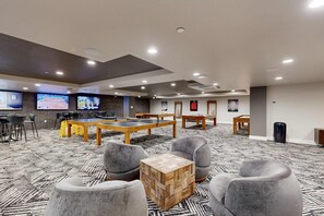 Un hall d’hôtel avec une table de ping-pong, des fauteuils gris, une table basse en bois et un grand écran diffusant un match de tennis.
