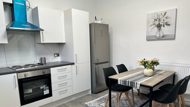 A modern kitchen with white cabinets, a black countertop, a built-in oven, a stainless steel refrigerator, and a dining area with a table and chairs. - Modern 3bedroom Home in Bury Premium Mattresses Blackout Blinds Ensuite M (Bury)