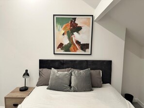 Ein Schlafzimmer mit Bett, Kissen, einem Nachttisch, einer Lampe und einem gerahmten abstrakten Gemälde an der Wand.