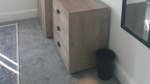 A room with a wardrobe, a small chest of drawers, a black trash bin, and a large mirror on the wall. - Modern 3bedroom Home in Bury Premium Mattresses Blackout Blinds Ensuite M (Bury)