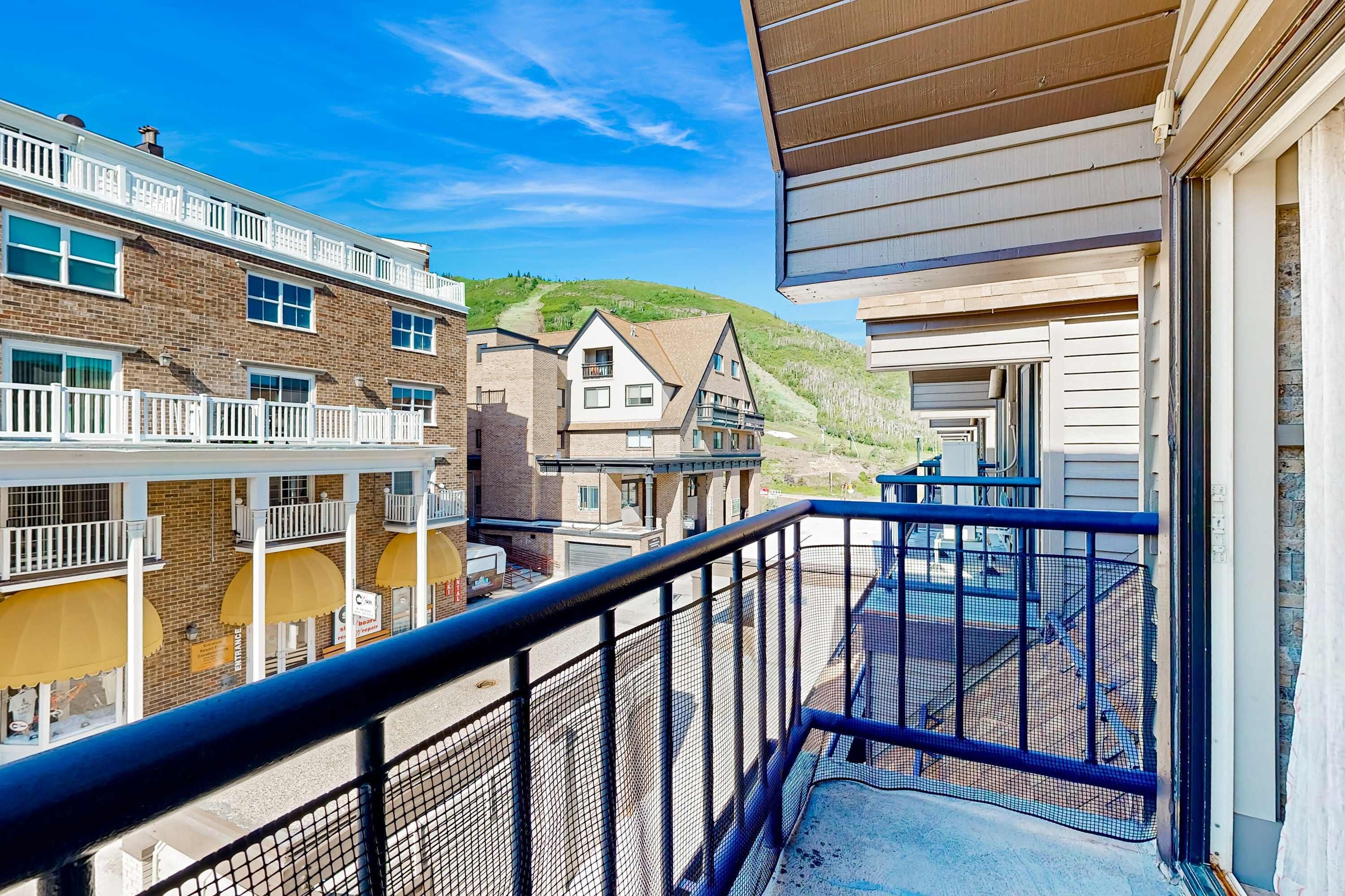 A balcony with a metal railing overlooking a street with buildings, a clear sky, and a hill in the background.