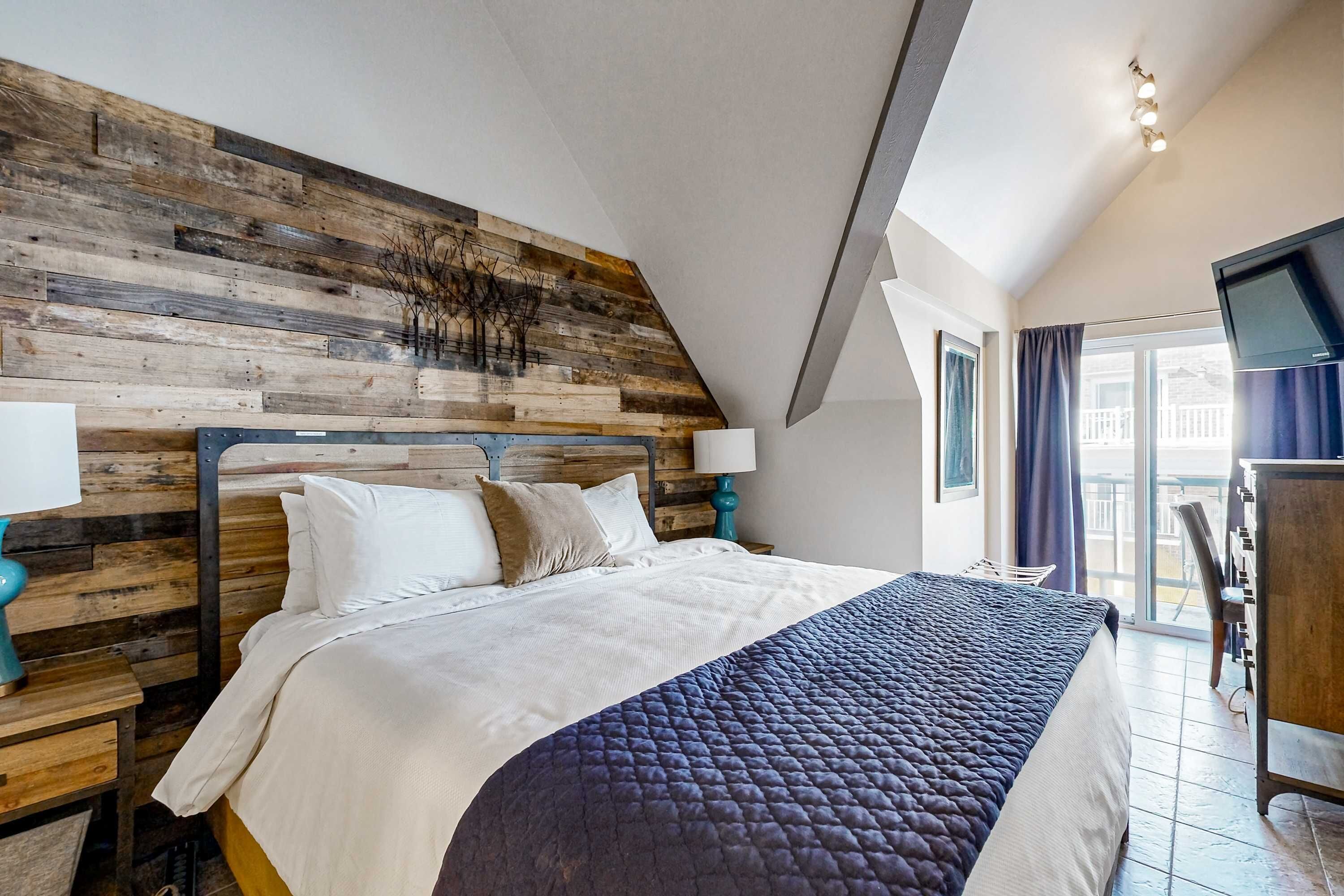Une chambre à coucher avec une tête de lit en bois, un lit avec une literie blanche et bleue, une table de chevet avec une lampe, et un téléviseur fixé au mur.