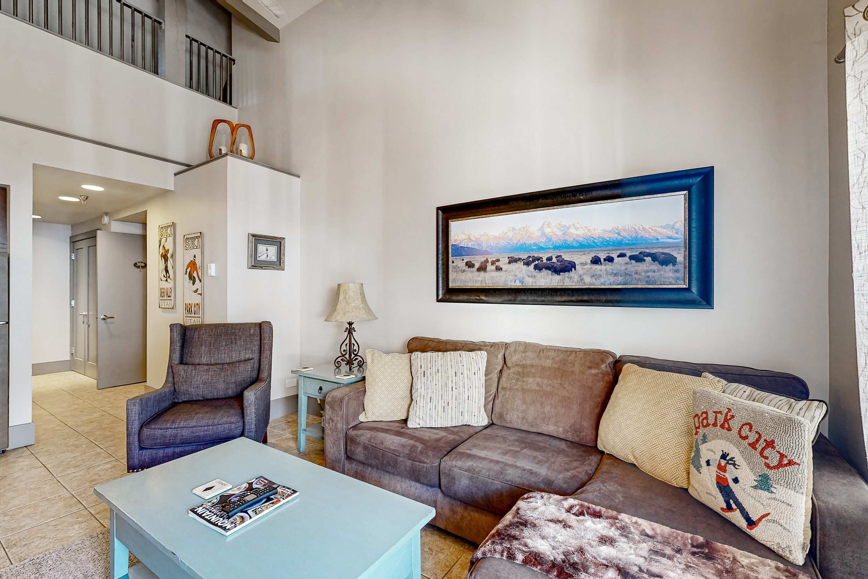 A living room with a sofa, armchair, and coffee table, a framed landscape painting, and a door leading to another room.