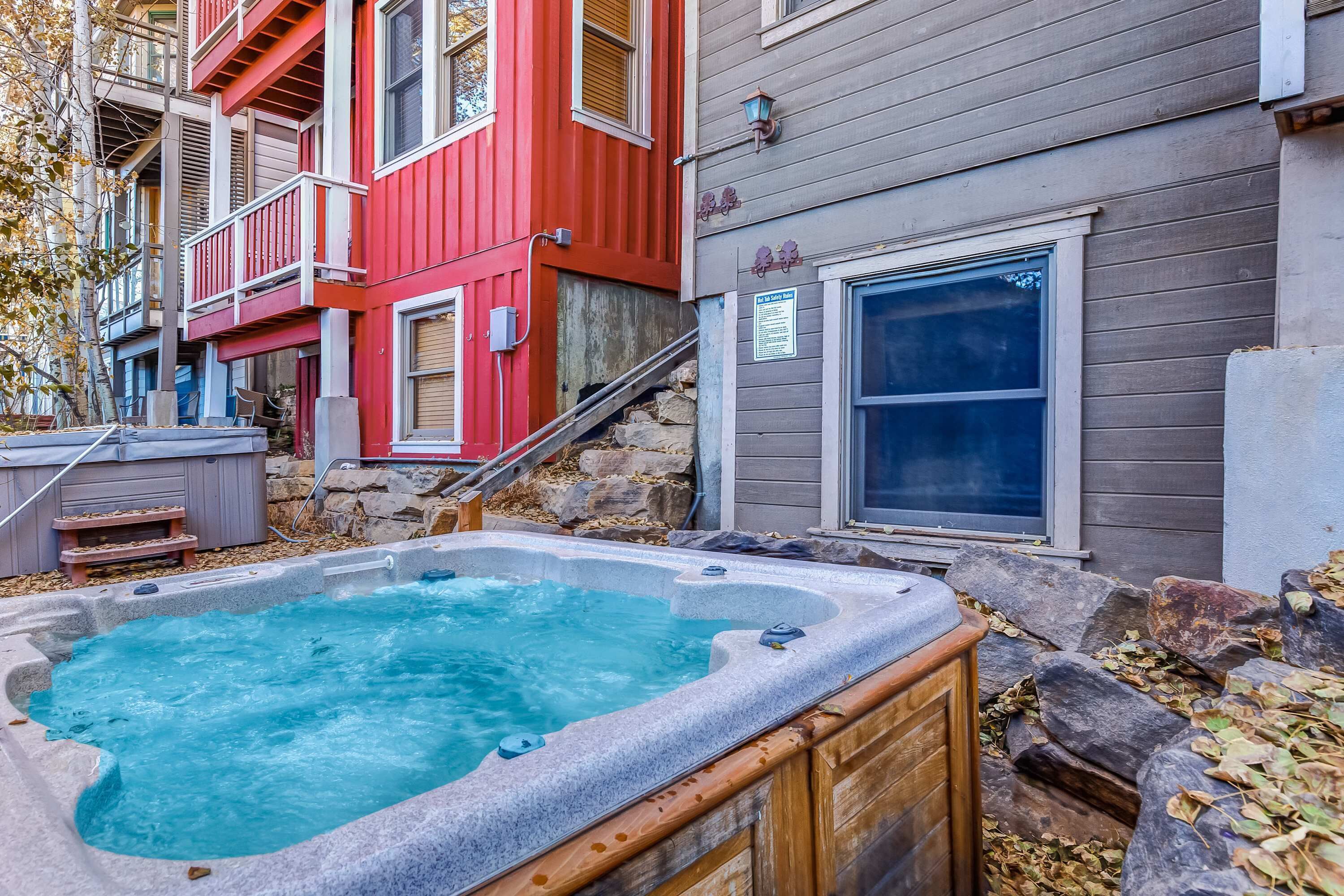 A hot tub in a backyard with a wooden deck, a stone wall, and a red building in the background.