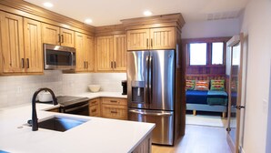 A kitchen with wooden cabinets, stainless steel appliances, a white countertop, and a view into a bedroom with a bed and colorful pillows. - Modern mountain escape with wonderful views, pool, hot tub-minutes to skiing (Park City)