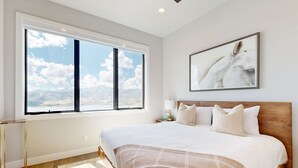 Een slaapkamer met een groot raam, een bed met witte lakens, een houten hoofdeinde en een lijstwerk met kunst aan de muur.