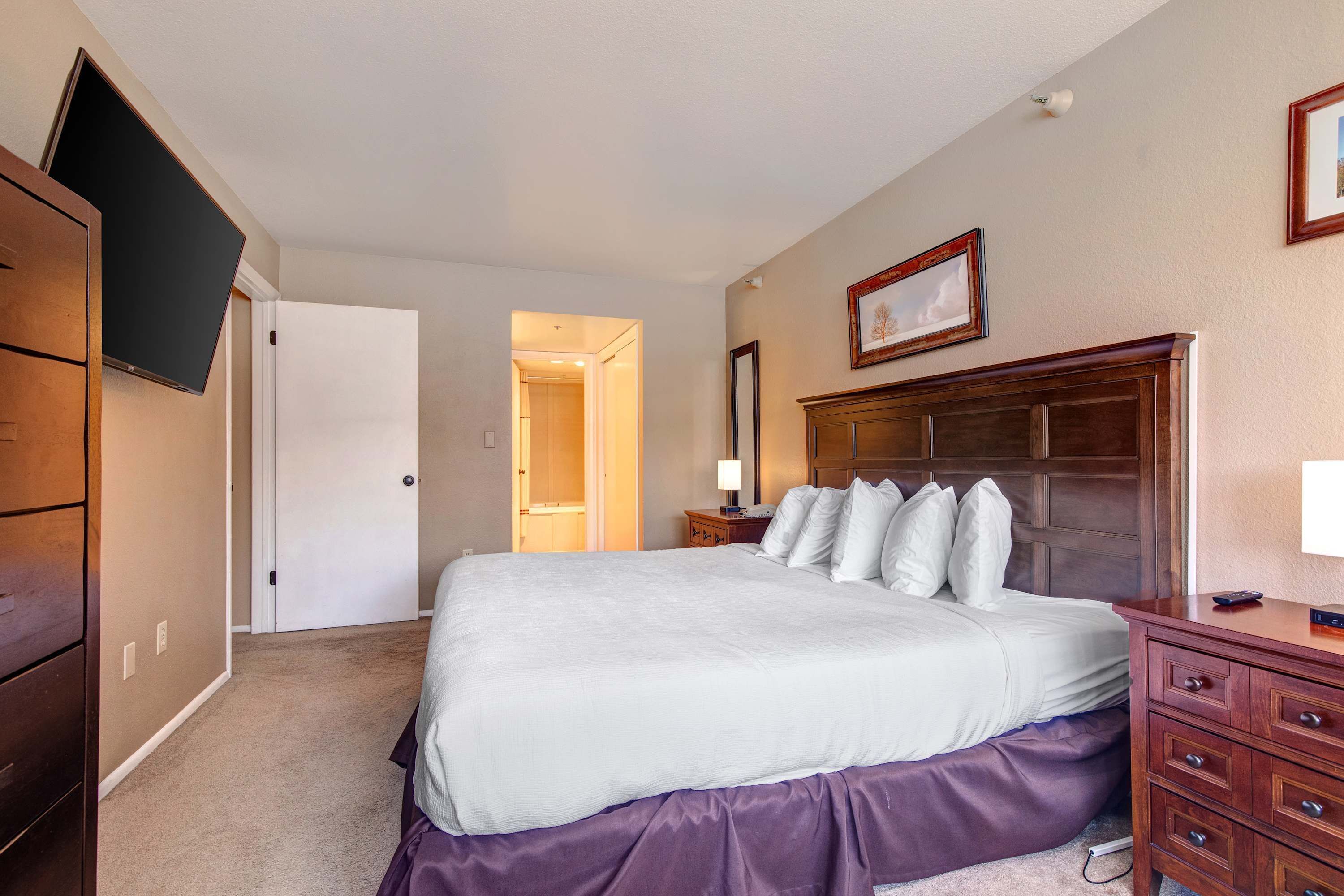 Une chambre à coucher comprenant un lit, une commode, un tél éviseur fixé au mur et une porte donnant sur une autre pièce.