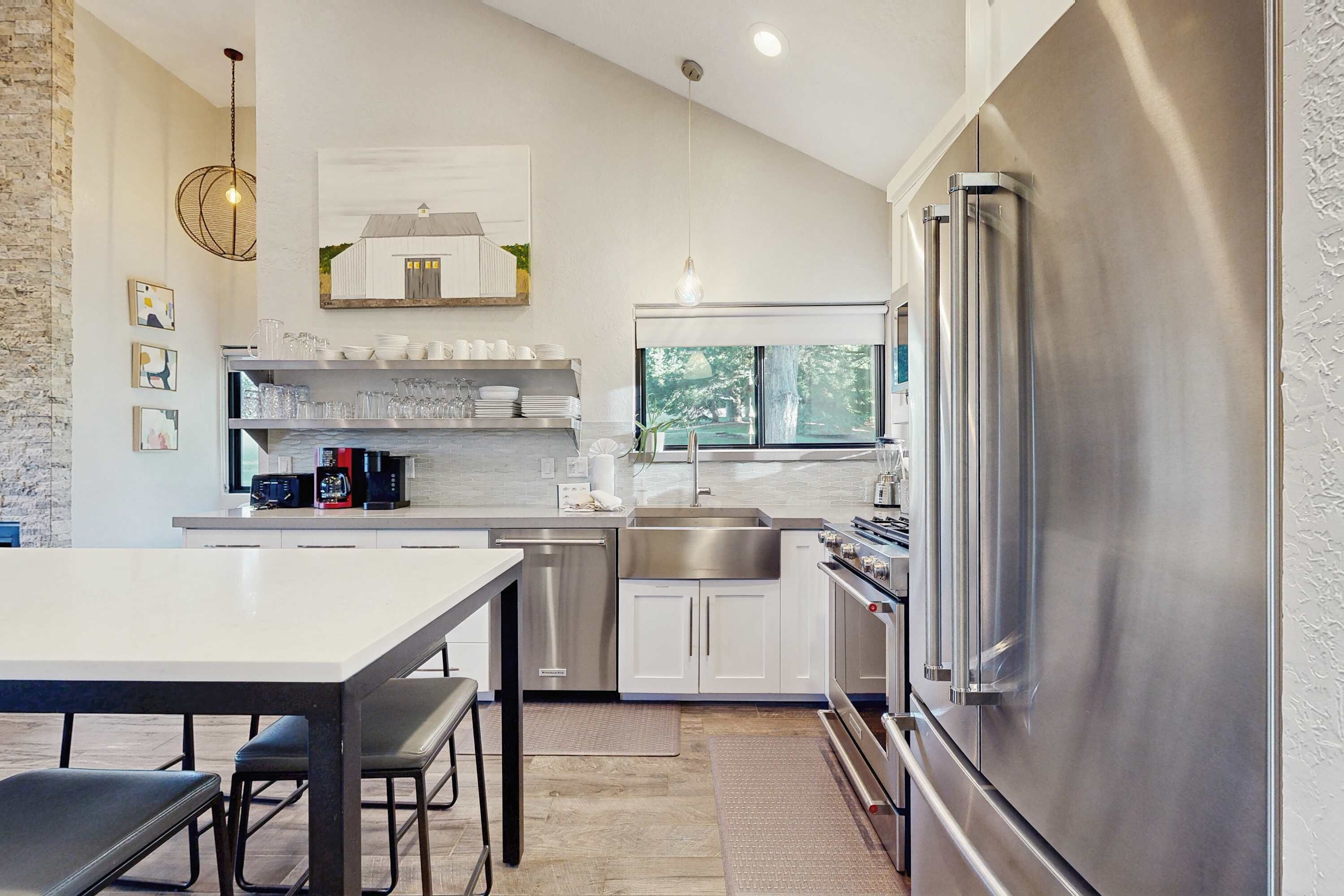 A modern kitchen with stainless steel appliances, a dining area with a white table and bar stools, and a window providing natural light.