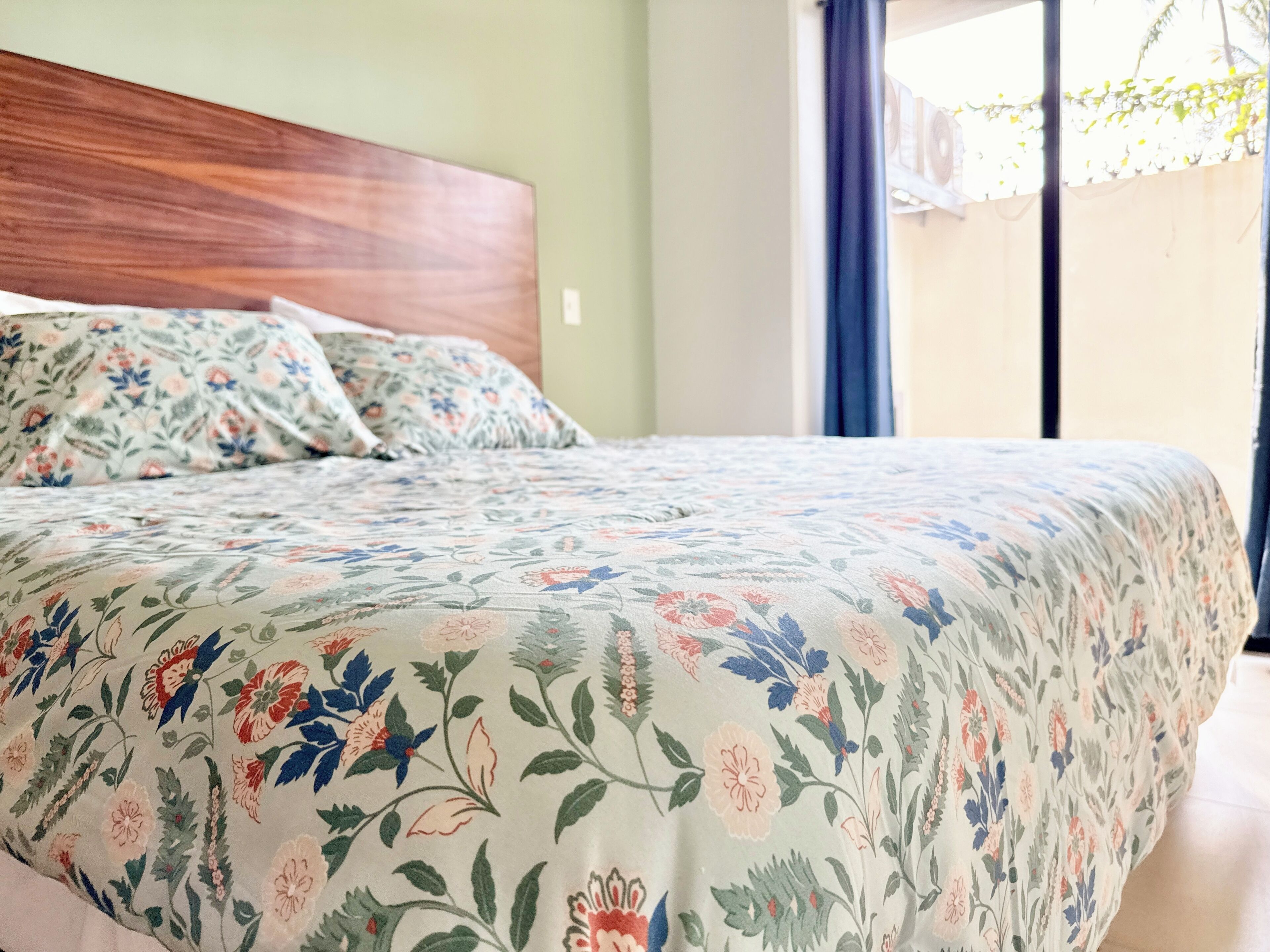 Una cama con una colcha estampada con motivos florales, un cabecero de madera y vistas a un balcón con barandilla y plantas.